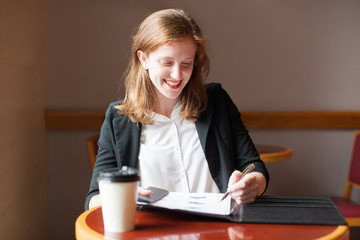 Portrait of happy young Caucasian businesswoman sitting at table with takeaway coffee and working...