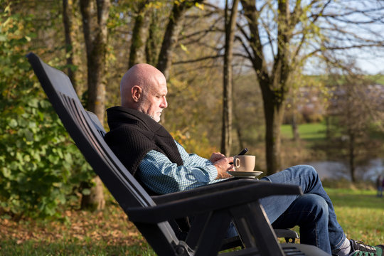 Senior man relaxing in chair outside with mobile phone and cup of coffee