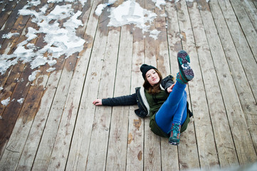 Young girl wear on long green sweatshirt, jeans and black headwear sitting on wooden floor with snow.