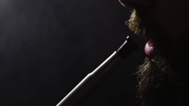young bearded man in glasses smokes a hookah and blow out smoke closeup on black background in slow motion in 4k