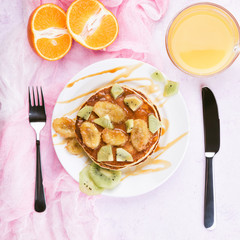 Homemade breakfast on a marble violet table. Stack of pancakes with a grilled bananas and a caramel. Glass of juice, orange, kiwi slices.Healthy breakfast concept, flat lay, top view