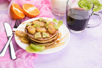 Homemade breakfast on a marble violet table. Stack of pancakes with a grilled bananas and a caramel. Glass of juice, orange, kiwi slices.Healthy breakfast concept, flat lay, top view