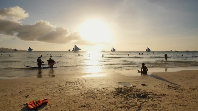 Beach on the island of Boracay in the rays of sunset. Holidaymakers on the beach. Sailboats on the water. Philippine Tropics.