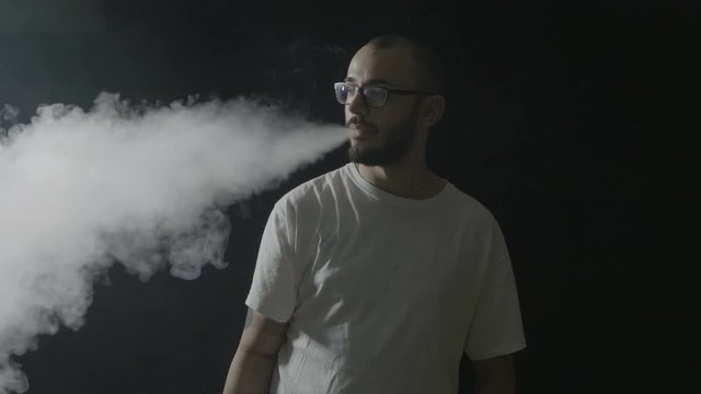 Teenager boy vaping having fun inhaling from a vaporizer and exhaling the smoke towards the camera on a dark background