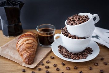 Espresso shot, coffee beans in the cup, coffee percolator and croissant on wooden table with black background