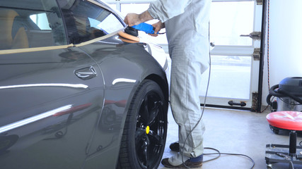 A man in a special suit polishes a gray car body, a tool for polishing cars, into a workshop.