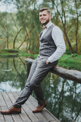 Handsome elegant bearded groom in checkered suit is standing on the bridge in the forest. Stylish wedding grooms photo in brown leather shoes. Rustic hipster portrait.