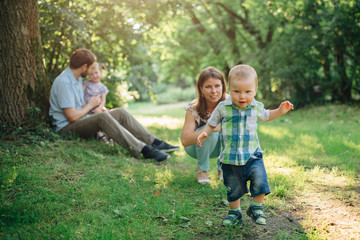 Fototapeta premium Cute little baby boy learning to walk in the park. Happy mother watching at her laughing son.