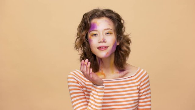 Portrait of beautiful female artist 20s with multicolored makeup blowing air kiss on camera and expressing romantic feelings, isolated over beige background. Body language