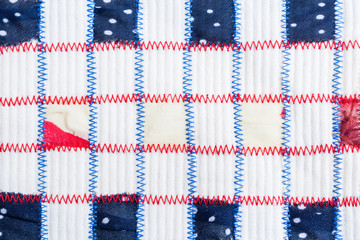 Square pieces of colored fabrics, sewn by red and blue zigzag seams on relief white fabric.