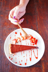 A piece of chocolate cake with strawberries lies on a white plate, sprinkled with jam. A small dessert fork lies side by side on a table with a wooden backdrop. Closeup view with copy space and hand