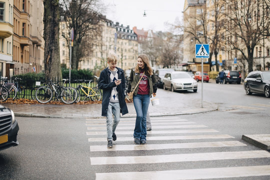 Smiling friends talking and looking at mobile phone while walking on zebra crossing in city