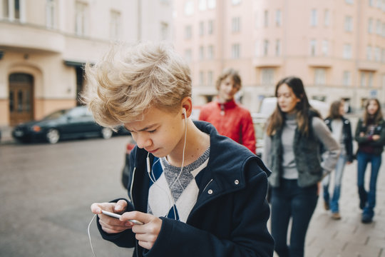 Teenage boy using mobile phone while walking with friends on sidewalk in city