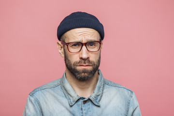 Obraz premium Grumpy bearded man with offended expression, being dissatisfied with something, frowns face, wears spectacles, hat and shirt, isolated over pink background. Displeasure and negativity concept