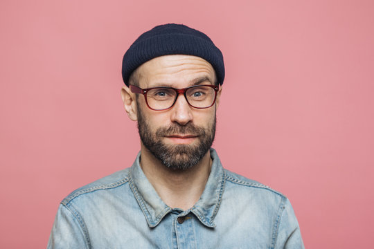 Portrait of handsome bearded man looks with pleased surprised expression, wears glasses and black hat, isolated over pink studio background. Middle aged unshaven male expresses bewilderment.