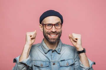 Happy bearded male with blue eyes and cheerful expression clenches fists, feels to be winner, demonstrates positiveness, isolated over pink background. Excited emotional young man poses indoor