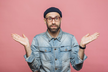 Photo of attractive bearded male wears fashionable clothing, shrugs shoulders in bewilderment, makes serious decision, isolated over pink background. Confused hesitant unshaven stylish man