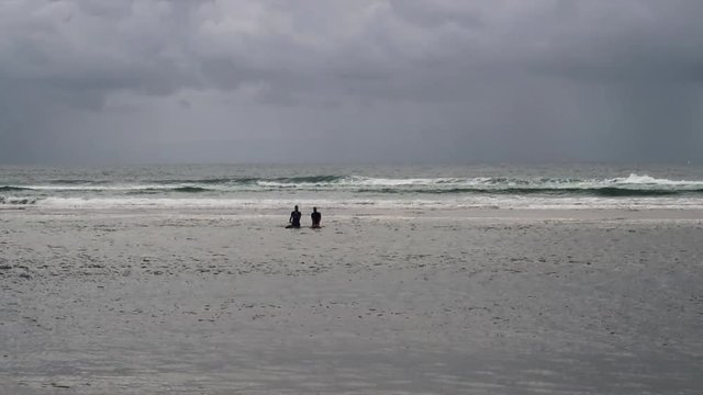 An Eye Level Long Shot of two Surfers in silhouette, walking through the water away from the Surf Break pushing their Surfboards. The white caps of the surf are highlighted against a stormy sky