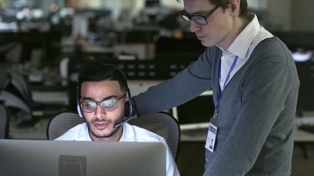PAN of male manager in glasses explaining something to Arab telemarketing agent in headset working on computer; name on badge is used in fictitious manner