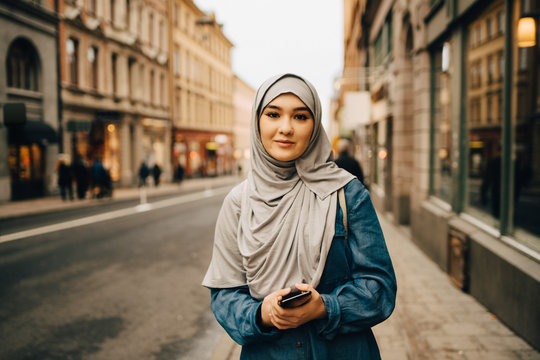 Portrait of confident young woman wearing hijab standing with mobile phone on sidewalk in city