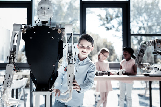 Cannot be cooler. Intellectual schoolboy smiling cheerfully while standing next to a human like robot and enjoying his time in a workshop.