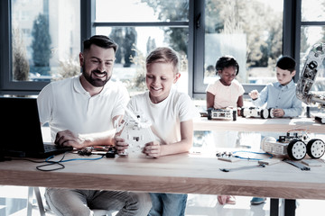 Examining the results of hard work. Selective focus on an extremely happy teacher grinning broadly while looking at a new robot of his preteen student while both sitting in a workshop.