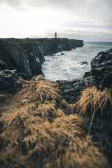 Orange lighthouse standing near a cliff with brown grass in the foreground and waves crashing against the rocks in the North of Iceland