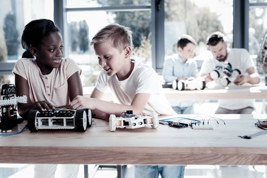 I like your idea. Beautiful African American girl looking at her best mate with a cheerful smile on her face while both sitting in a workshop and constructing robotic machines together.