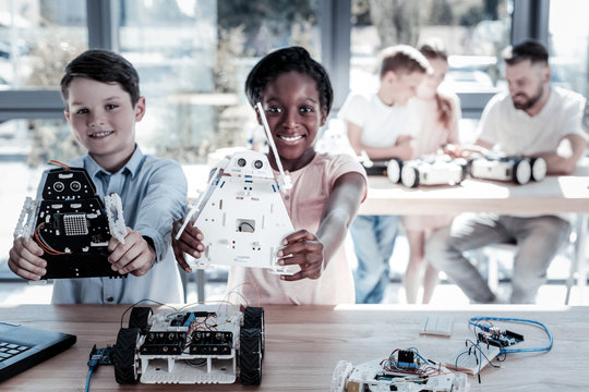 We had so much fun constructing them. Selective focus on impressive robots held by proud girl and boy looking into the camera with smile on their faces in a workshop.