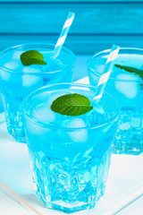 Blue cocktail with ice and mint in glasses on a white wooden board on a blue table.
