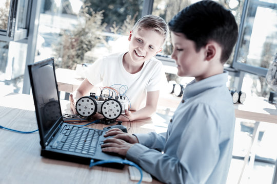 Are you done. Selective focus on a blond boy smiling while holding a newly constructed robotic vehicle and looking at his friend programming on a laptop.