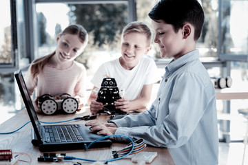 Intelligent trio. Selective focus on brown haired little boy sitting next to his friends and working on a program for their robots in workshop.