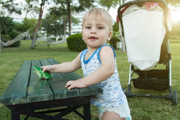 A small child in a diaper plays with a toy frog in the street, next to a bench. Green bench, lawn,...