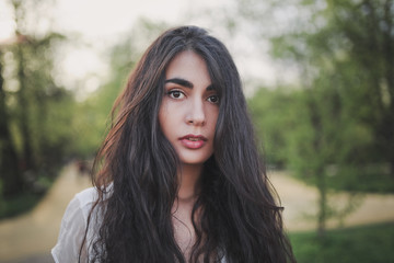 Obraz premium Lovely brooding brunette girl with long hair in white romantic dress in spring forest or park. The atmosphere of a warm sunny evening.