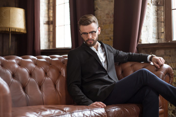 Handsome stylish confident bearded businessman in a smart suit and glasses sitting on a sofa in the...