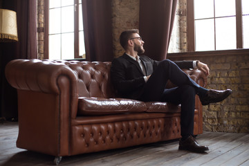 Handsome stylish confident bearded businessman in a smart suit and glasses sitting on a sofa in the...