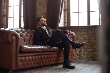 Handsome stylish confident bearded businessman in a smart suit and glasses sitting on a sofa in the...