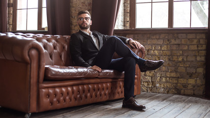 Handsome stylish confident bearded businessman in a smart suit and glasses sitting on a sofa in the...