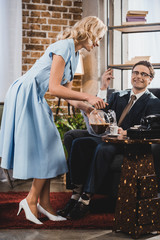 smiling woman pouring coffee to happy husband in suit and eyeglasses sitting in armchair, 1950s...