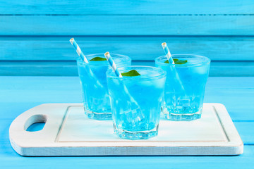 Blue cocktail with ice and mint in glasses on a white wooden board on a blue table.