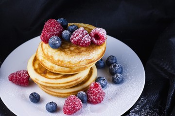 Stack of pancakes with raspberries and blueberries  coated with powdered sugar on white plate and black background