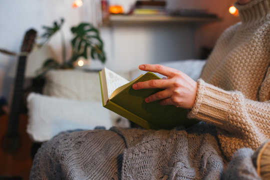 Woman Reading A Book, Sitting In A Cozy Chair With Kn It Plaid In Front Of A Fireplace, Winter Cozy Concept, Soft Toned Image.