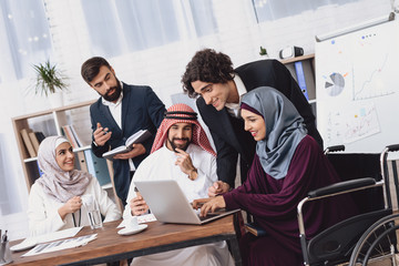 Disabled arab woman in wheelchair working in office. Woman is sharing notes on laptop.