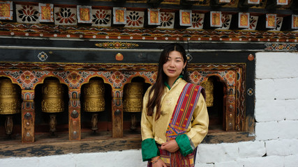 Bhutanese young woman facing the camera with a smile is wearing a kira (national dress) and standing in front of prayer wheels in monastery - obrazy, fototapety, plakaty