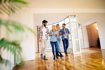 A group of young cheerful business people walking down the bright hallway while discussing and drinking coffee from cups.