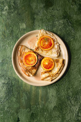 Homemade crepes pancakes served in ceramic plate with bloody oranges and rosemary syrup with sliced sicilian red oranges over green texture background. Top view, space