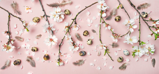 Easter holiday background. Flat-lay of tender Spring almond blossom flowers on branches, feathers, quail eggs over light pink background, top view. Greeting card concept
