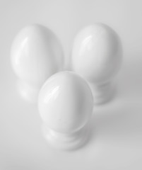Top view of three white decorative ceramic eggs on stands or in egg-cups on white blurred background. Easter religious Christian symbol. Monochrome image
