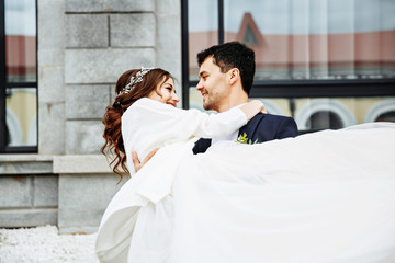the groom holds his bride in his arms and they dance near the building with beautiful windows