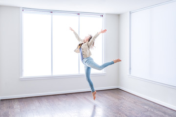 One young happy woman jumping up in empty modern new room with hardwood floors and large sunny windows in apartment